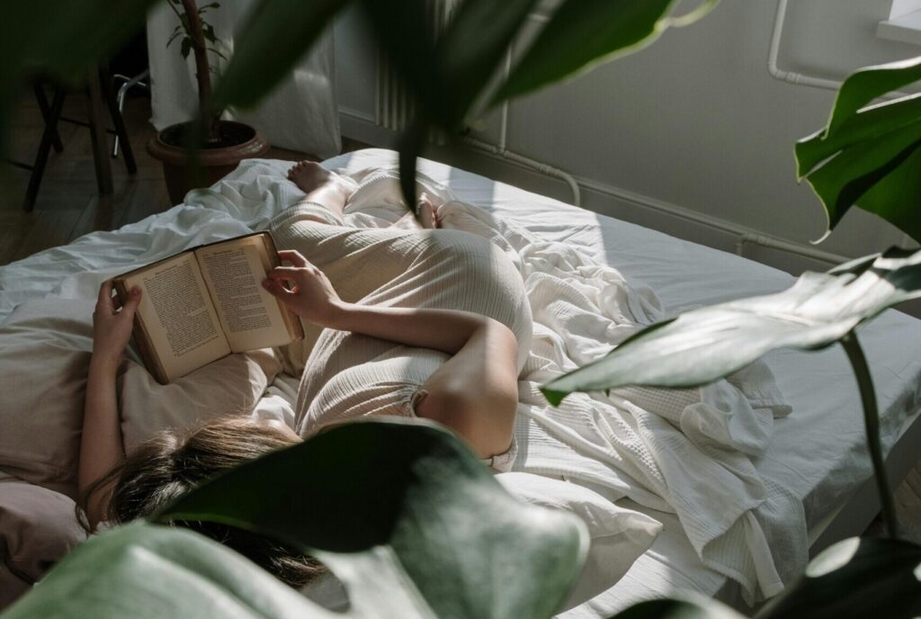 A woman lies in bed reading a book, surrounded by greenery and natural light.